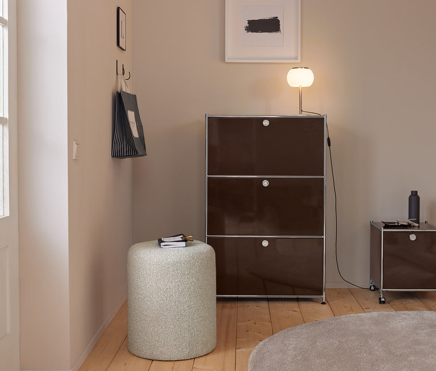 Entrée moderne avec murs clairs, parquet et tapis rond beige. Une grande armoire modulaire marron foncé avec une lampe lumineuse se tient à côté d'une armoire plus petite à roulettes. Un pouf cylindrique clair avec un carnet est au premier plan. Un sac fourre-tout rayé est accroché à des crochets sur le mur gauche.