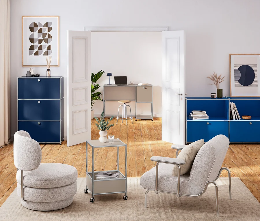 Deux fauteuils clairs sur un tapis. À gauche, une commode en métal « CN3 » bleu foncé, à droite un buffet en métal « CN3 » bleu gentiane. Entre eux, une table basse en métal « CN3 » taupe. À l'arrière-plan, un bureau en métal « CN3 » taupe avec lampe de table LED et tabouret.