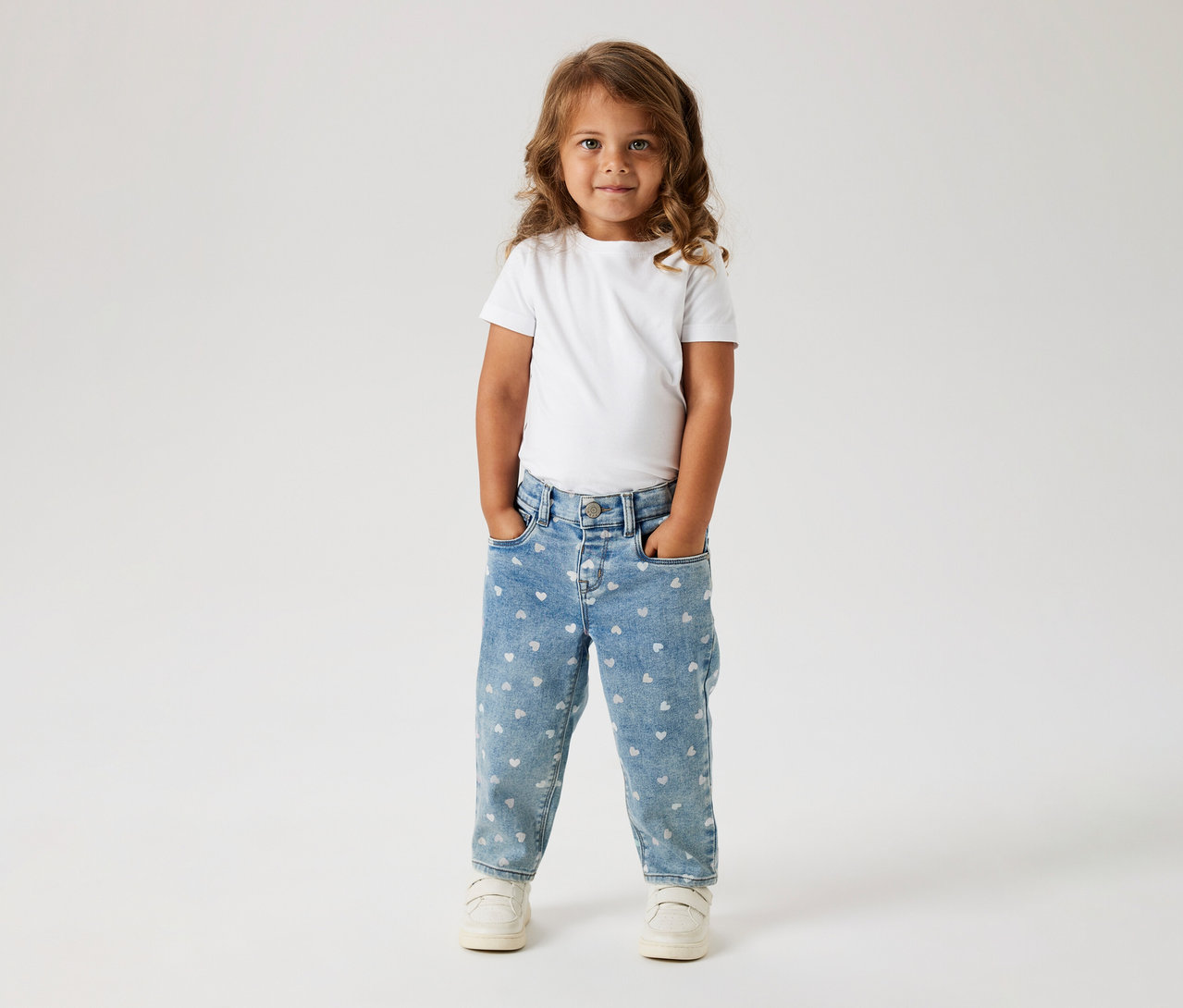 Un jeune enfant aux longs cheveux bruns ondulés se tient de face, portant un t-shirt blanc, un jean bleu clair à motifs cœurs blancs et des baskets blanches. Ses mains sont dans ses poches.