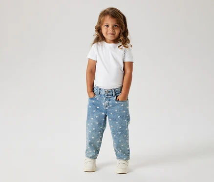 Un jeune enfant aux longs cheveux bruns ondulés se tient de face, portant un t-shirt blanc, un jean bleu clair à motifs cœurs blancs et des baskets blanches. Ses mains sont dans ses poches.
