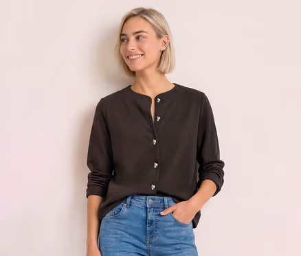 Jeune femme aux cheveux blonds porte un cardigan Street One marron foncé avec des boutons argentés et un jean, souriant sur le côté.