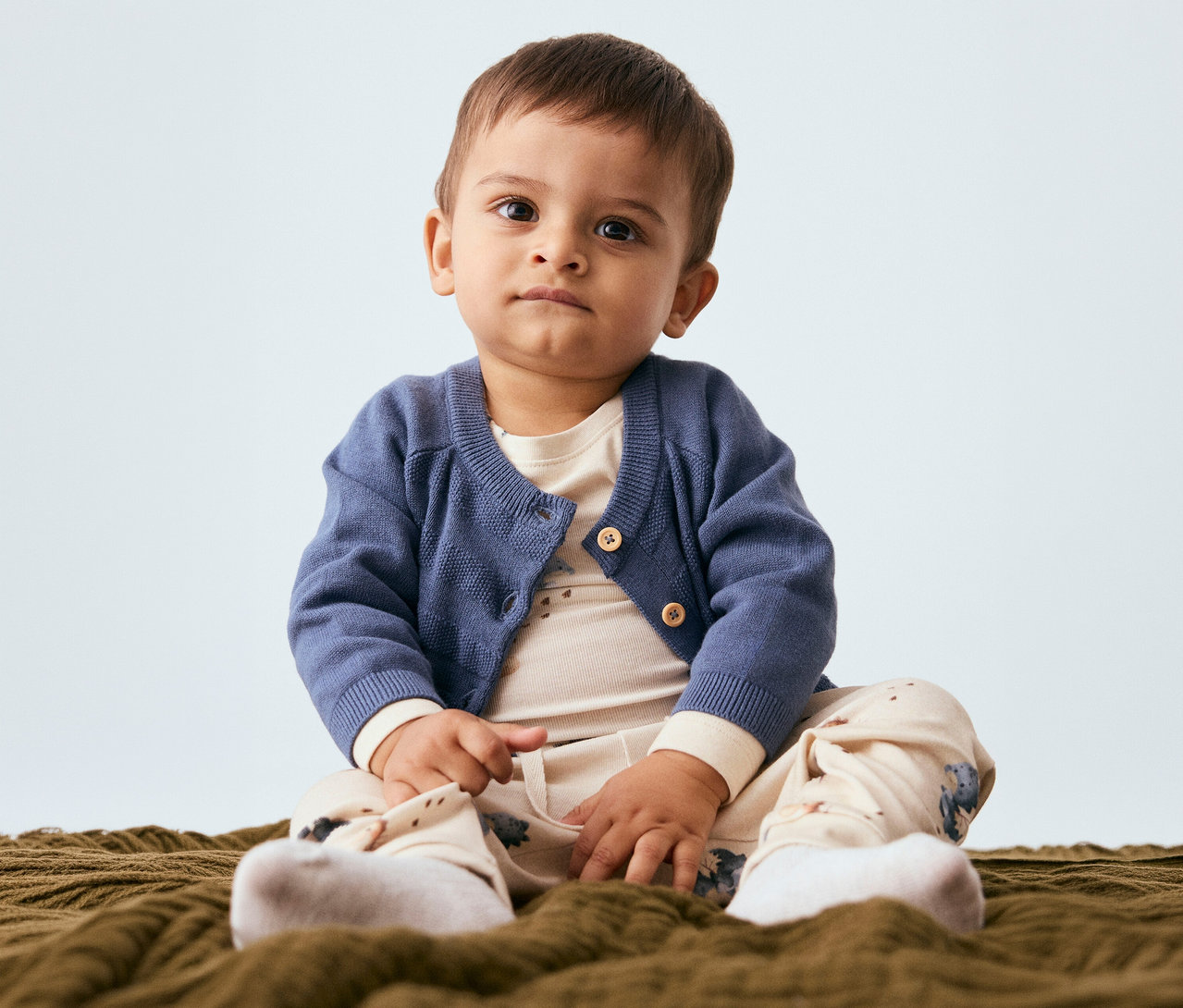 Un bébé est assis sur une couverture, portant un cardigan bleu, un haut beige clair, un pantalon à motifs et des chaussettes blanches. Il regarde l'appareil photo.