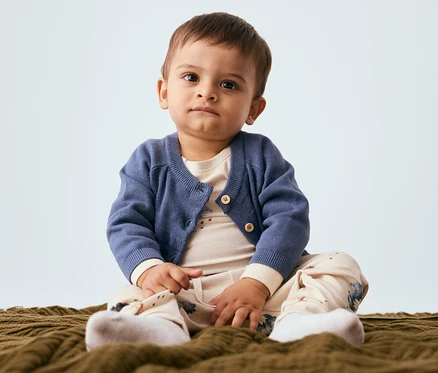 Un bébé est assis sur une couverture, portant un cardigan bleu, un haut beige clair, un pantalon à motifs et des chaussettes blanches. Il regarde l'appareil photo.