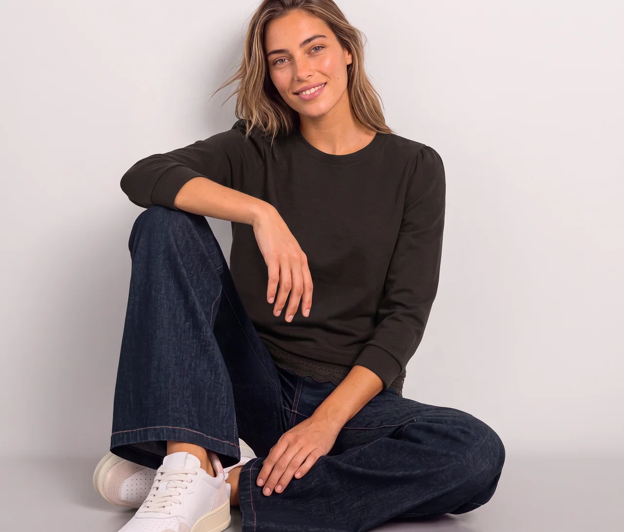 Une femme souriante est assise par terre, vêtue d'un T-shirt en jersey 3/4 Street One marron à manches légèrement bouffantes, d'un jean large et de baskets claires.