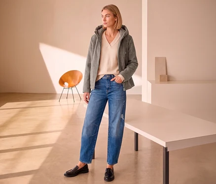 Une femme aux cheveux blonds porte une veste de mi-saison Street One grise, un pull col en V beige, un jean bleu et des mocassins noirs.