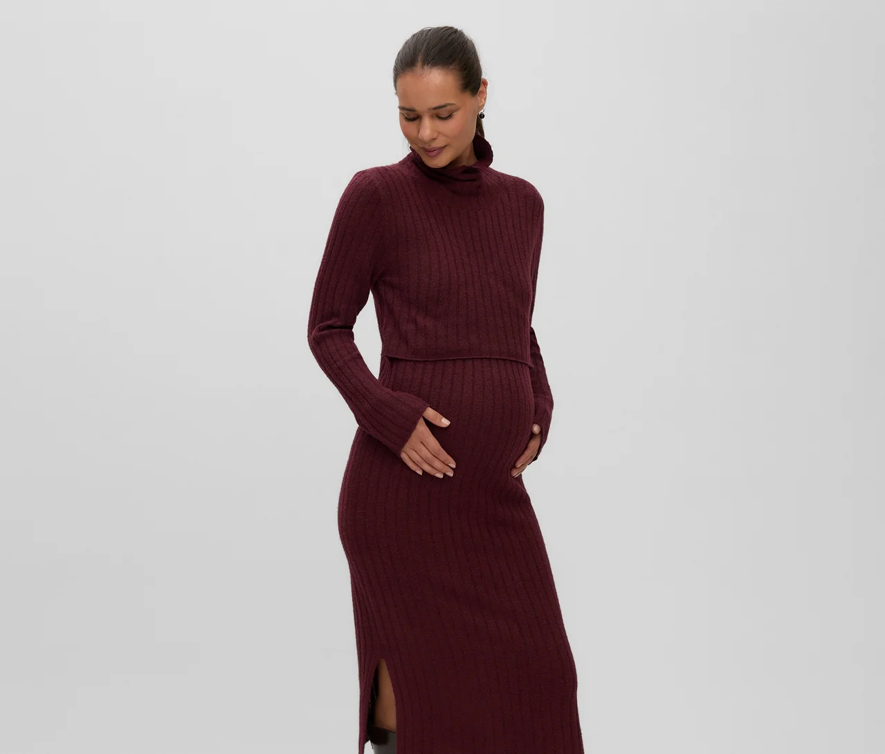 Femme enceinte portant une robe mi-longue en maille côtelée bordeaux MAMALICIOUS « SUNIVA » avec col montant et fente latérale, tenant son ventre.
