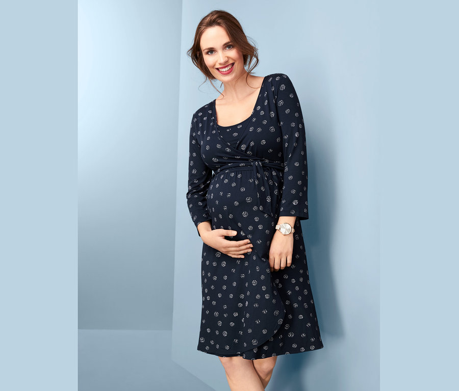 Une femme enceinte souriante tient son ventre et porte une robe de grossesse bleu marine à l’aspect cache-cœur avec un imprimé.