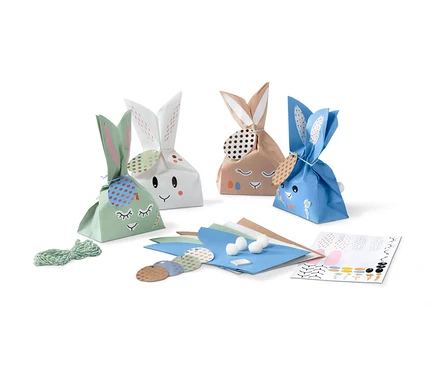 Quatre pochettes cadeau en forme de lapin avec un visage et des oreilles sont placées côte à côte. À côté d'eux se trouvent des matériaux de décoration.