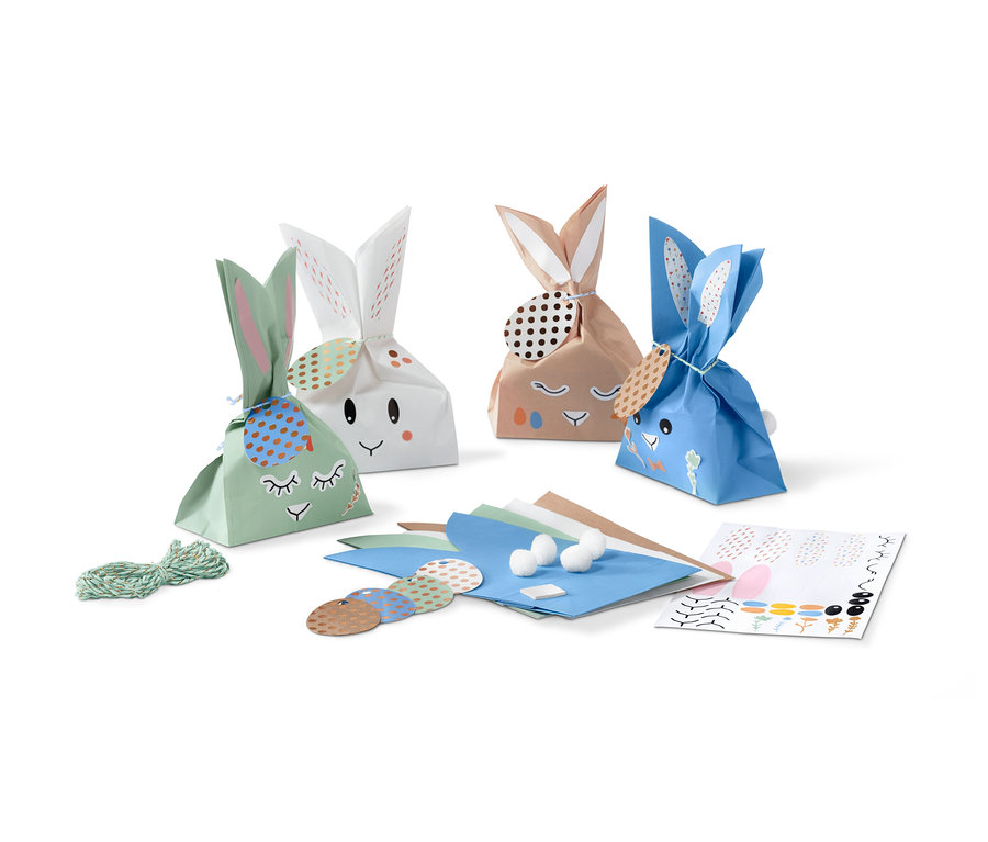 Quatre pochettes cadeau en forme de lapin avec un visage et des oreilles sont placées côte à côte. À côté d'eux se trouvent des matériaux de décoration.
