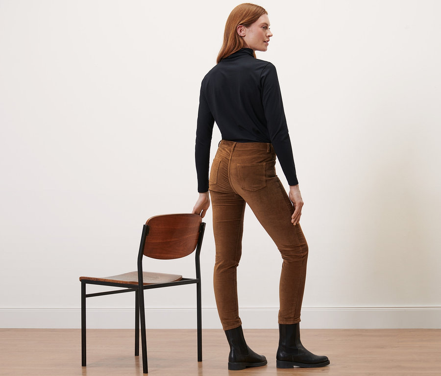 Une femme se tient de côté et porte un pantalon en velours cognac, des bottes noires et un col roulé.