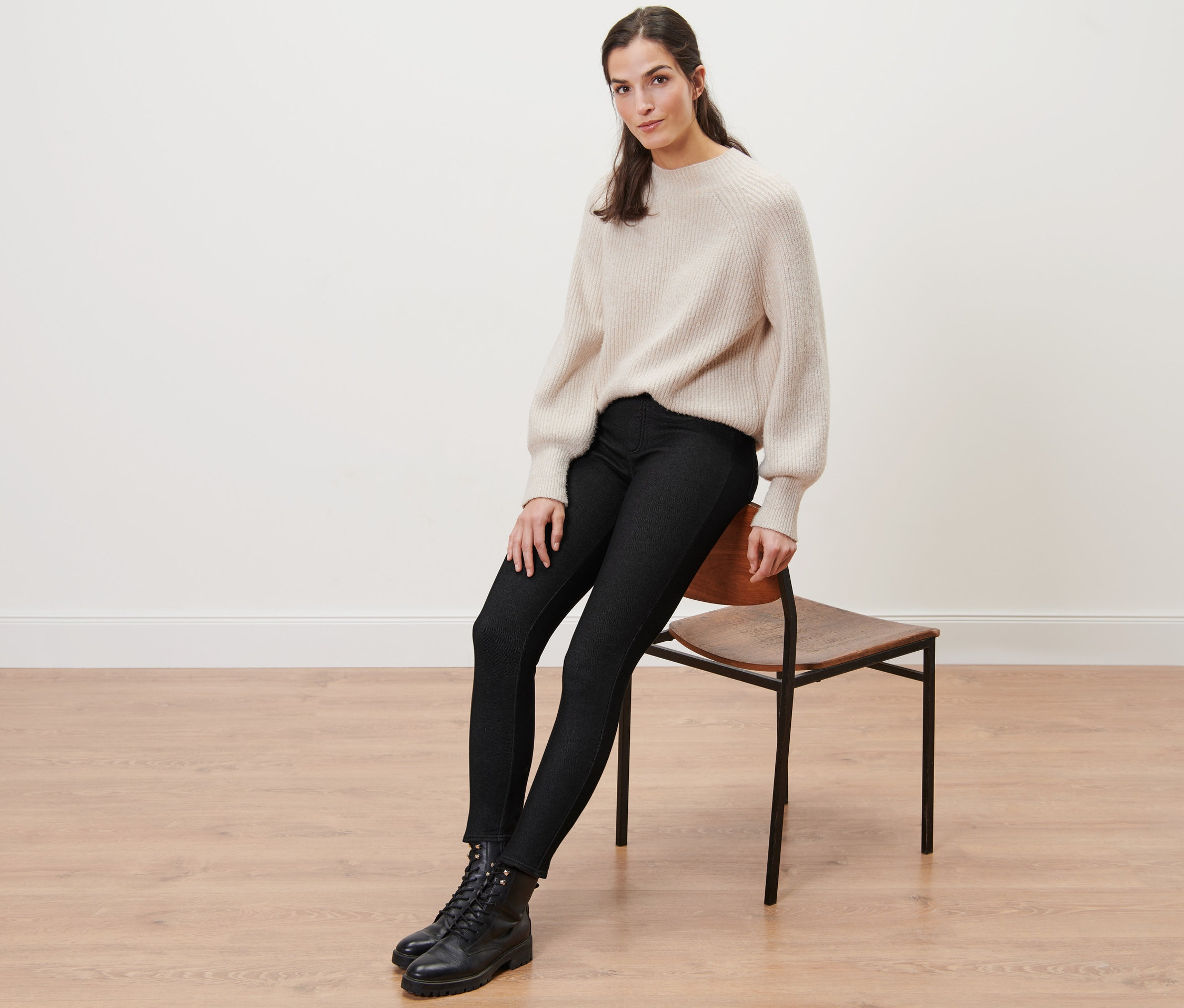 Une femme est assise sur une chaise et porte un legging doublé à l’aspect jean, un pull et des bottes.