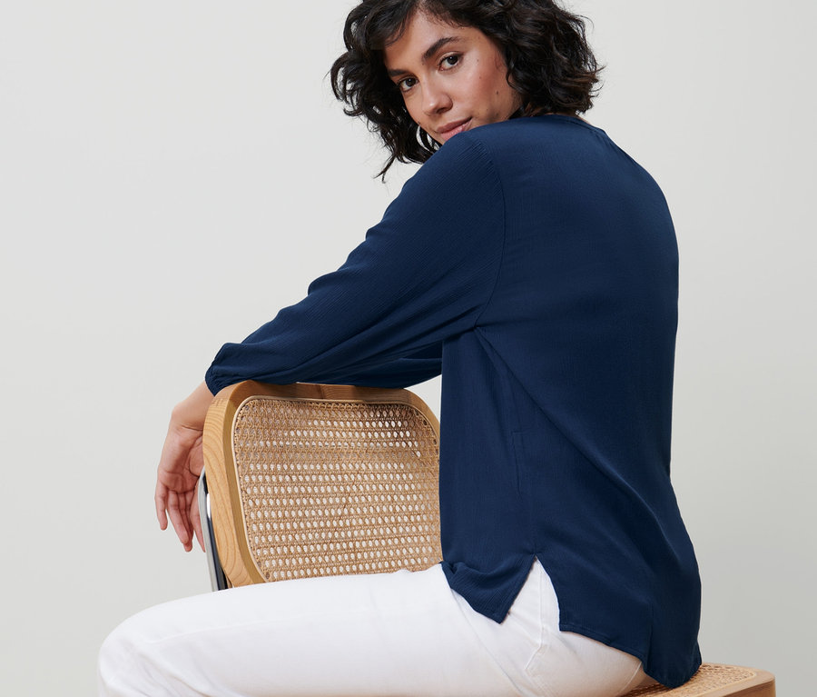 Une femme est assise de côté sur une chaise et porte un chemisier-tunique bleu.