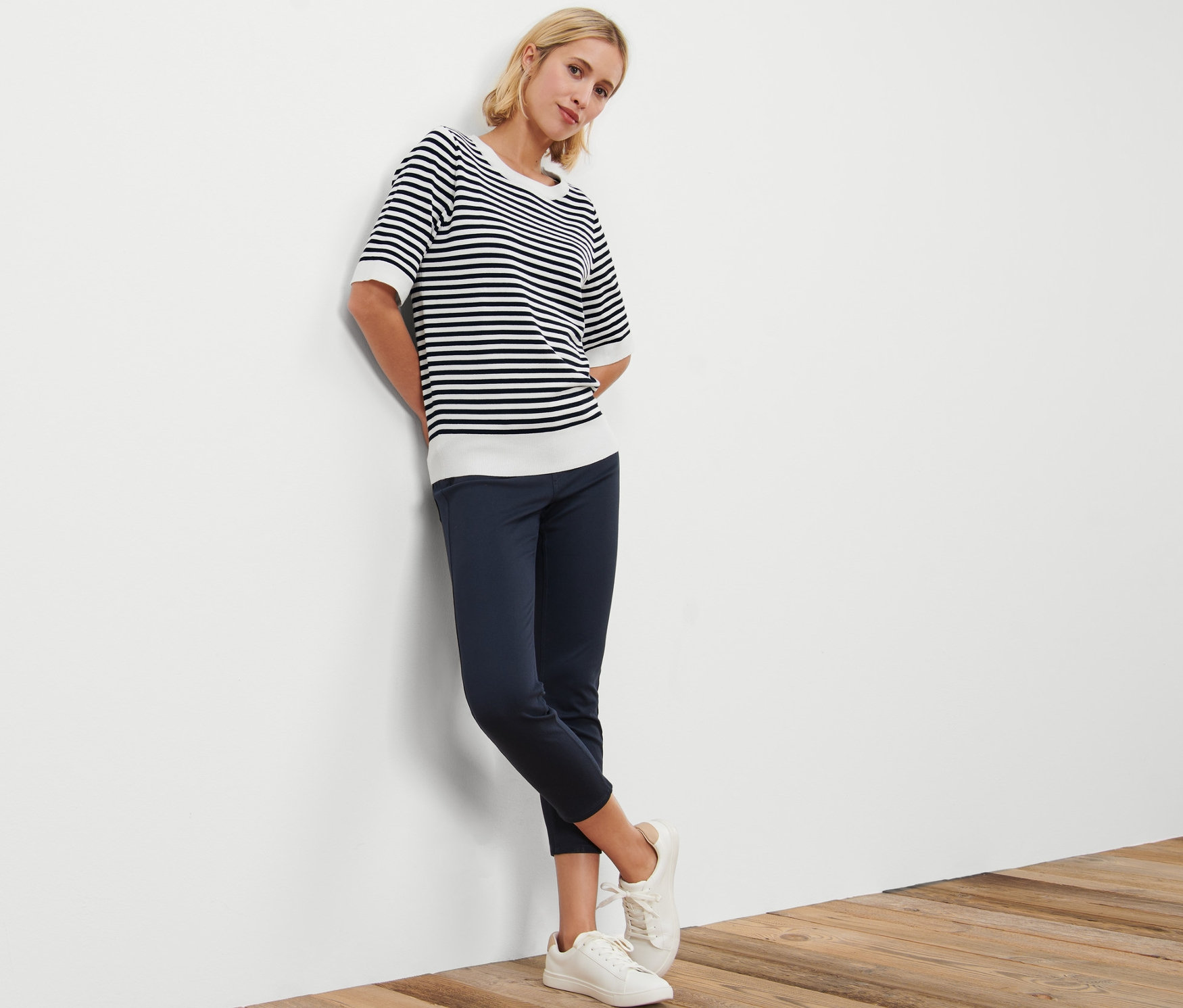 Femme appuyée contre un mur portant un pull rayé en maille fine, un pantalon bleu marine et des baskets blanches.