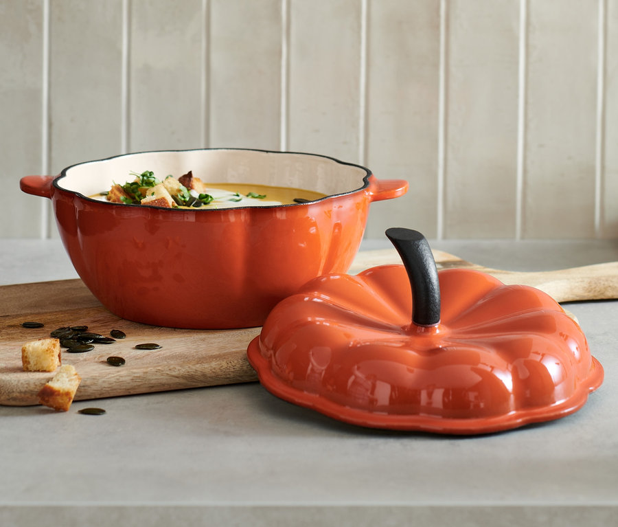 Un pot ouvert en fonte en forme de courge rempli de soupe avec des croûtons est posé sur une planche à découper en bois à côté du couvercle.