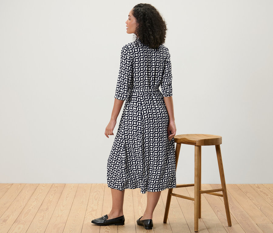 Une femme se tient à côté d'un tabouret en bois et porte une robe chemise imprimée.