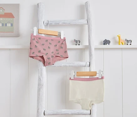 Deux culottes pour enfant suspendues à une échelle blanche. Une est rose avec un motif zébré, l'autre est couleur crème avec une bordure rose.