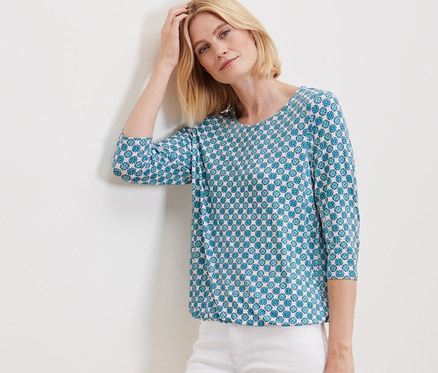 Femme posant contre un mur en blouse avec imprimé intégral et pantalon blanc.