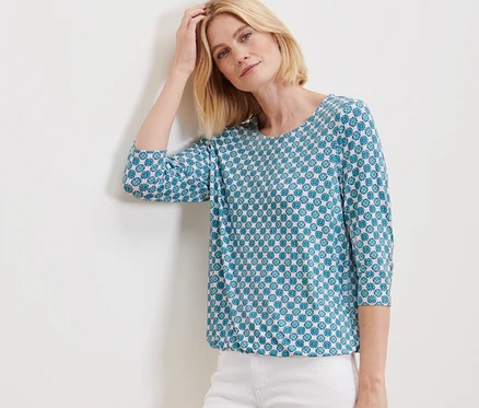 Femme posant contre un mur en blouse avec imprimé intégral et pantalon blanc.