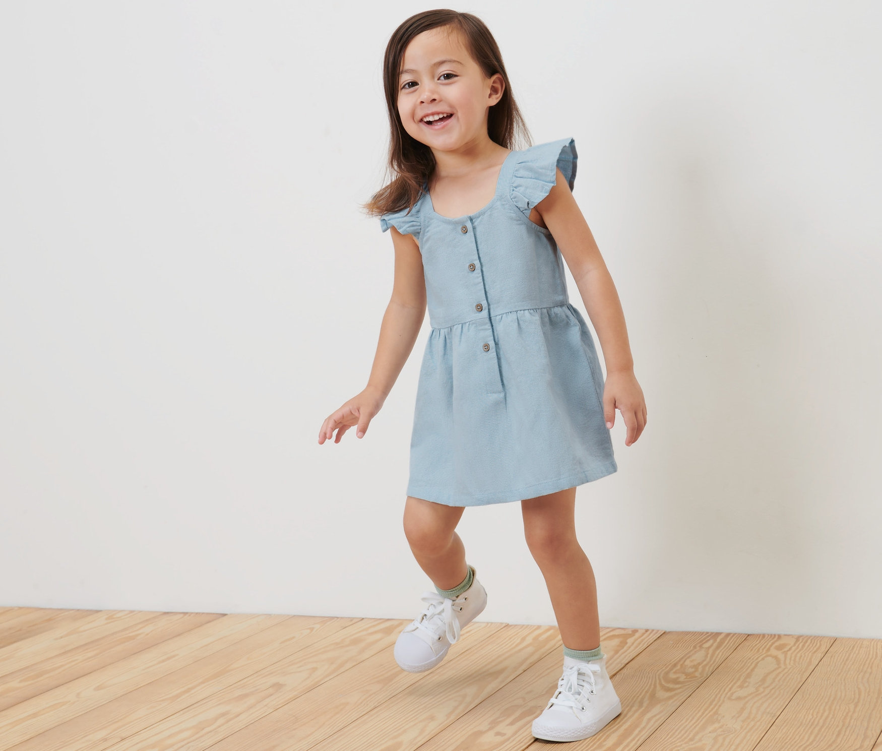Fillette souriante marchant sur un plancher en bois et portant une robe tisée pour enfant.