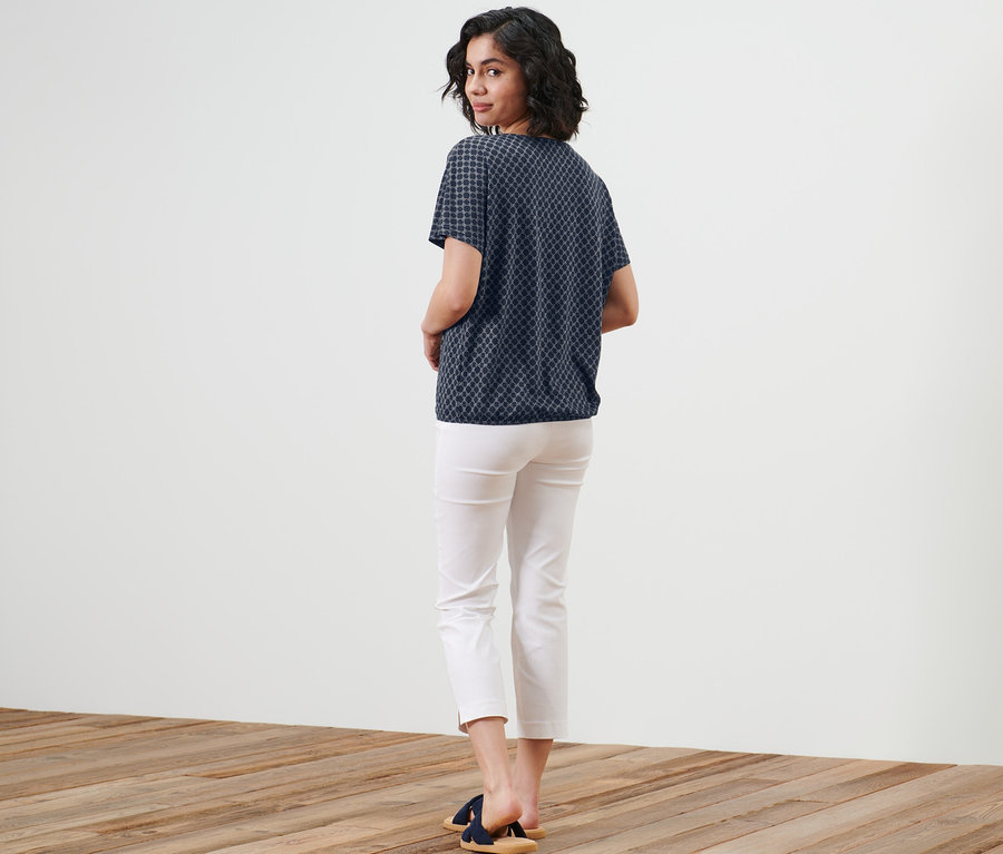 Une femme regarde par-dessus son épaule et porte une blouse imprimée, un pantalon blanc et des sandales.