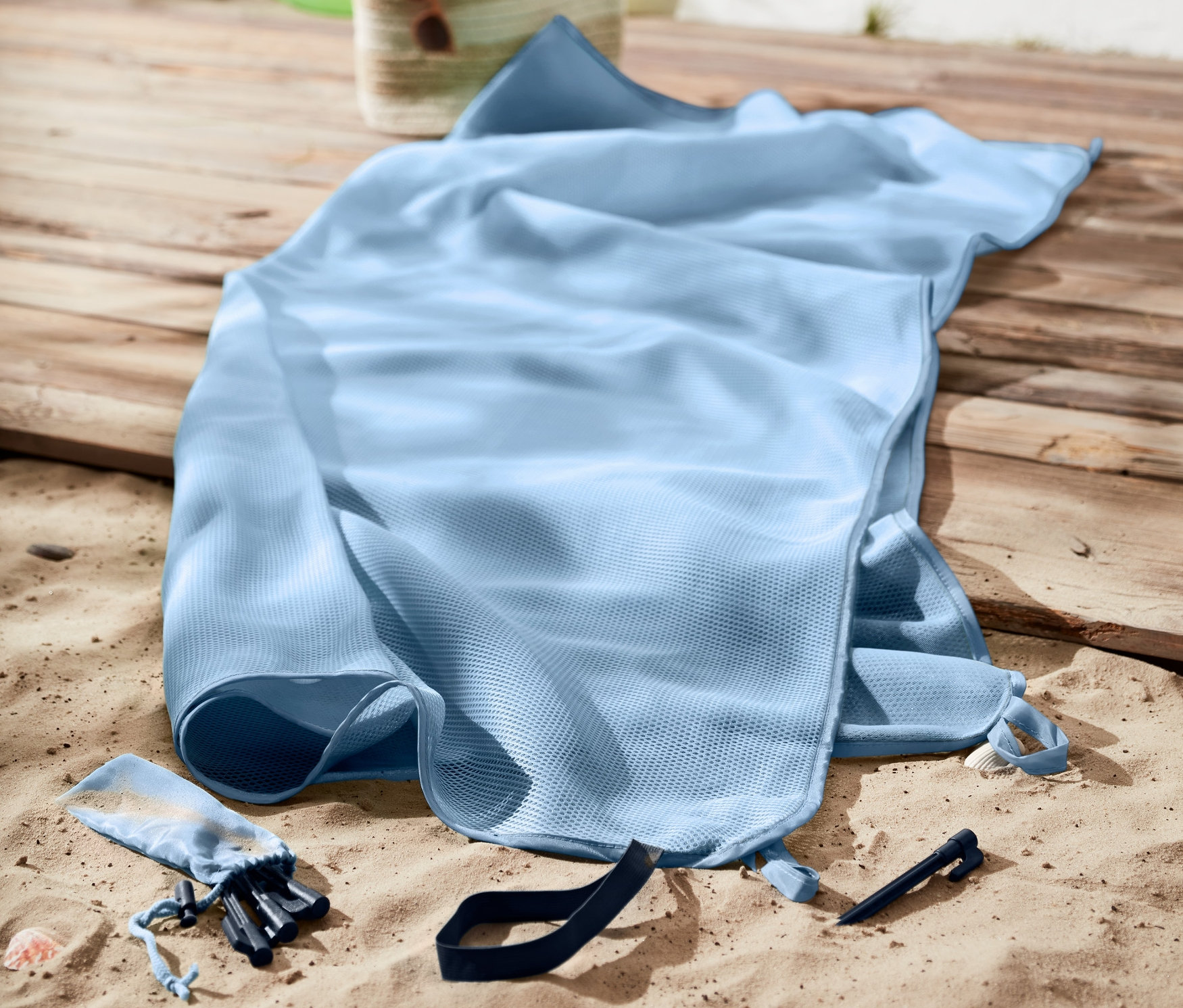 Une couverture de plage anti-sable bleu clair est posée sur le sable. Un anneau de natation vert et un sac sont visibles à l'arrière-plan.