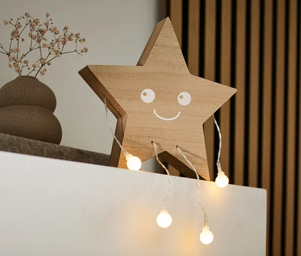 Sur une étagère blanche se trouvent une figurine décorative à LED « Étoile » en bois avec un visage souriant, un vase avec de petites fleurs et un livre.