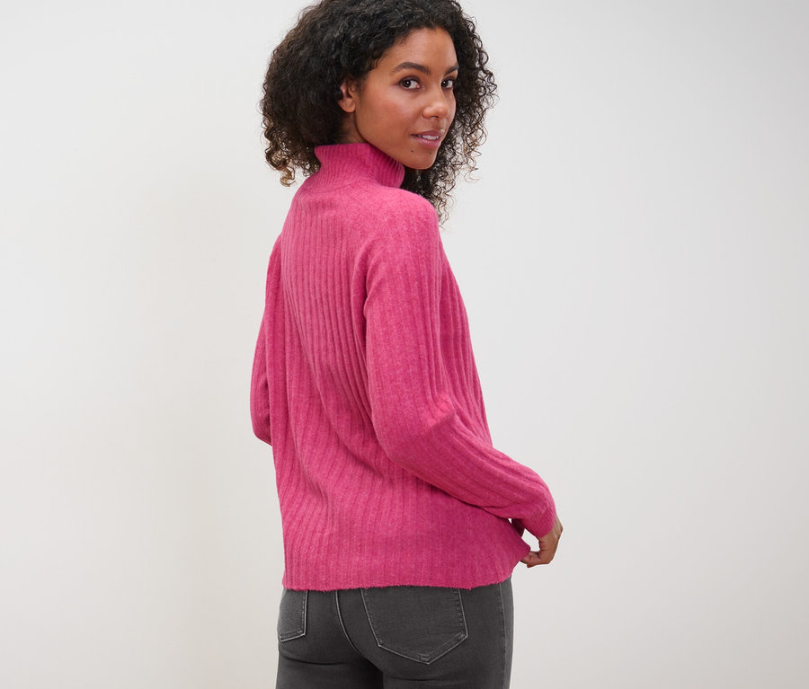 Femme regardant par-dessus son épaule portant un pull en maille côtelée à col montant et un jean gris.