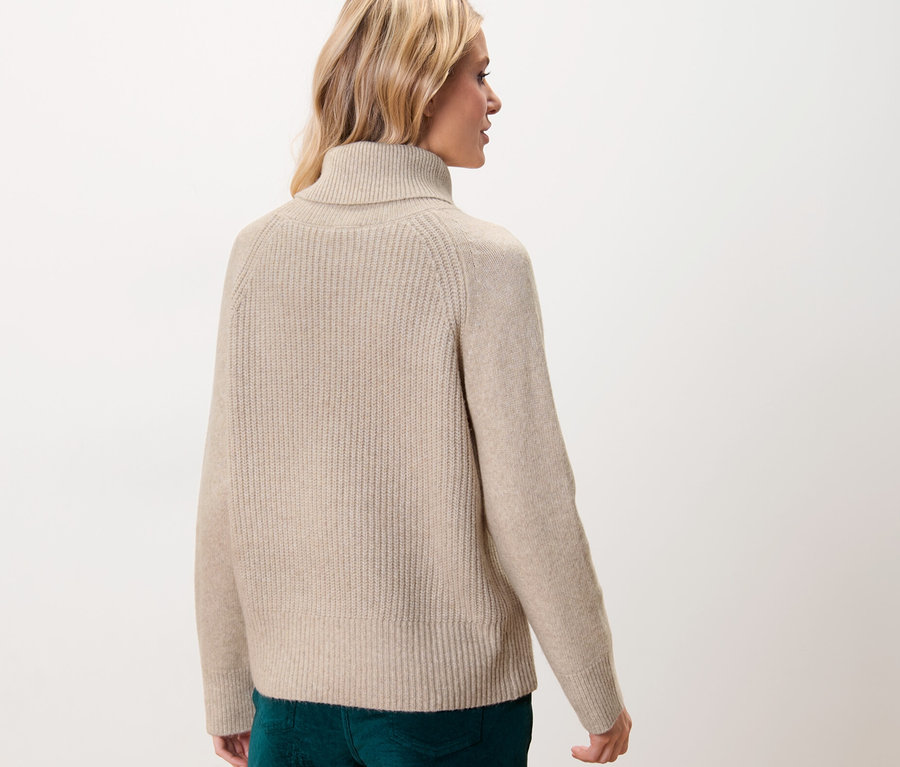 Une femme blonde, vue de dos, porte un pull à col roulé en grosse maille beige.
