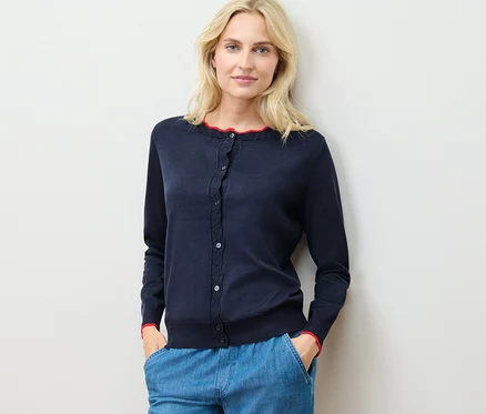 Une femme porte un cardigan à maille fine avec bord ondulé et un pantalon bleu, debout contre un mur.
