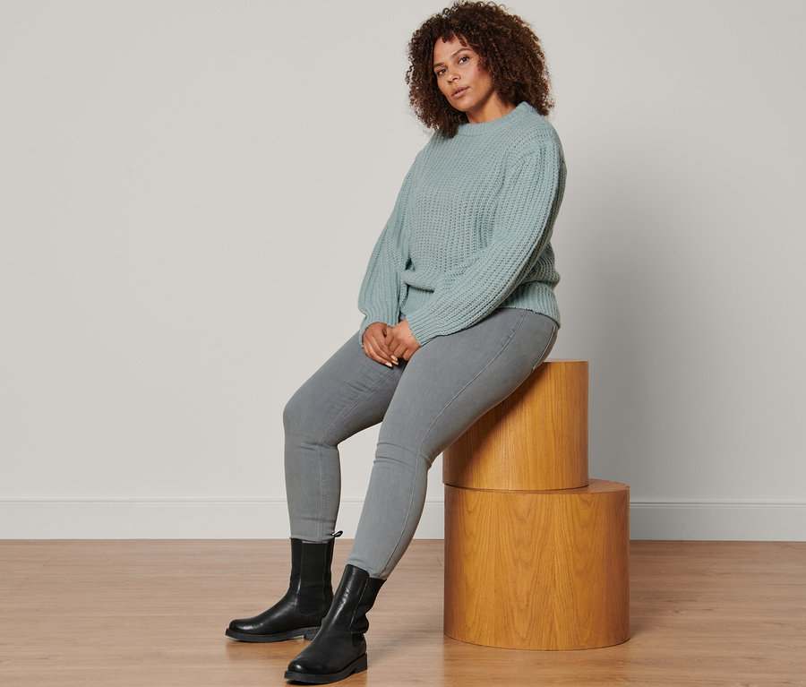 Une femme assise sur des socles en bois porte un pull en grosse maille bleu fumé avec de la laine, un tregging gris chiné et des bottines en cuir.