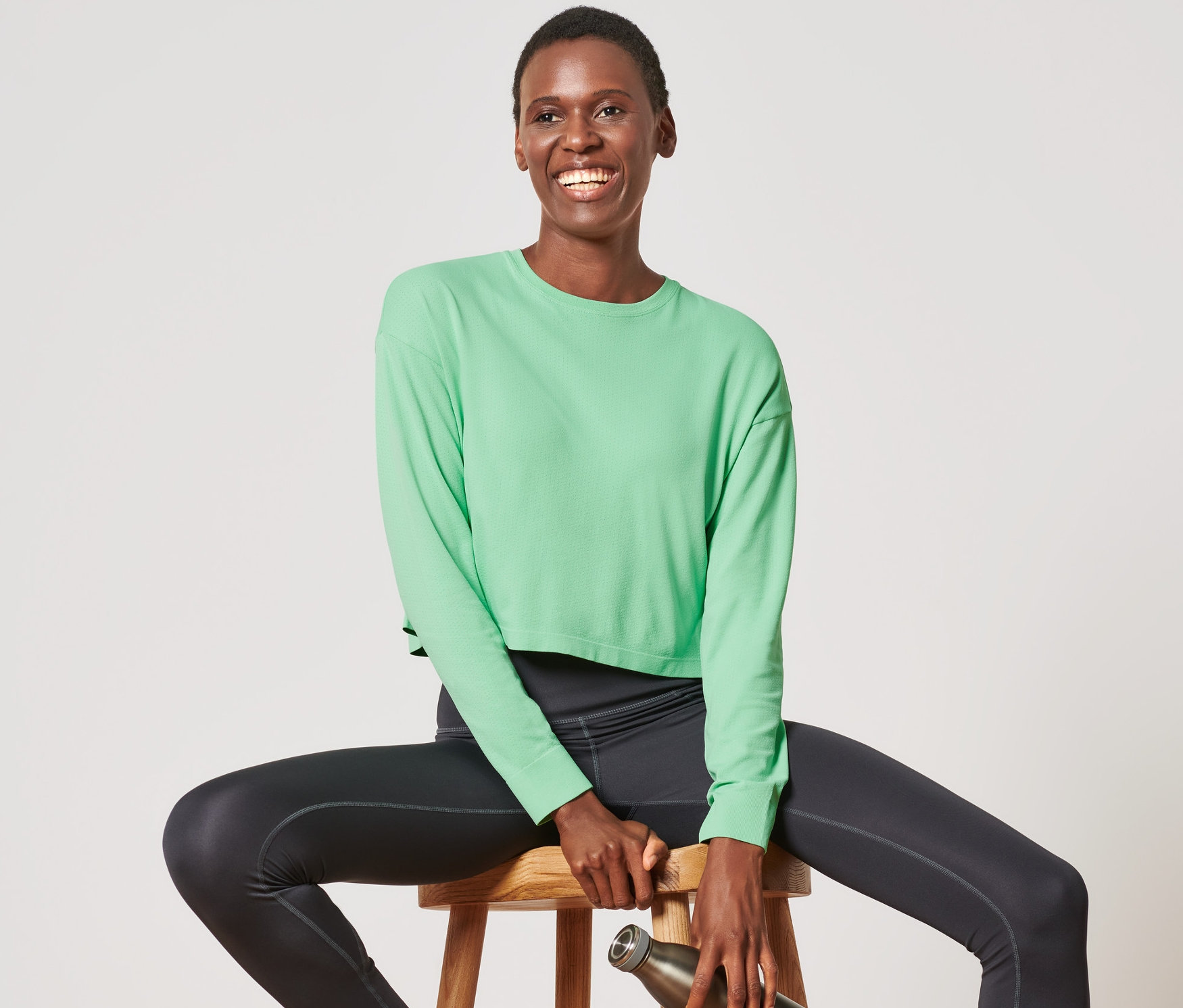 Une femme est assise sur un tabouret en bois, portant un t-shirt fonctionnel sans coutures vert clair, des leggings noirs et des baskets blanches. Elle tient une bouteille d'eau en métal dans sa main.