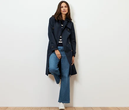 Une femme adossée à un mur portant un trench-coat bleu foncé.