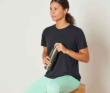Femme assise sur un tabouret en bois tenant une bouteille en métal, portant un t-shirt de sport anthracite.