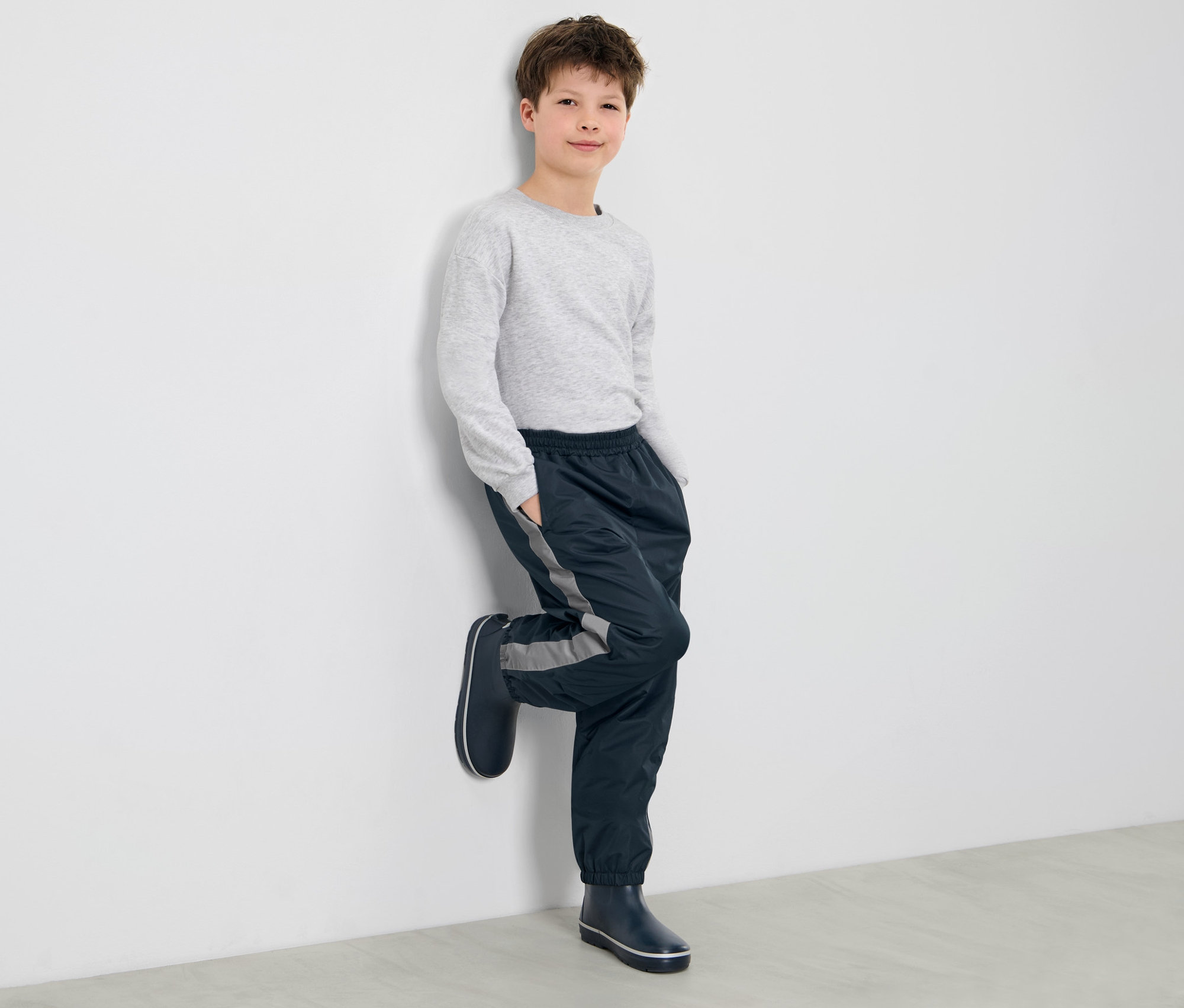 Un garçon appuyé contre un mur porte un pantalon de pluie pour enfant et des bottes en caoutchouc.