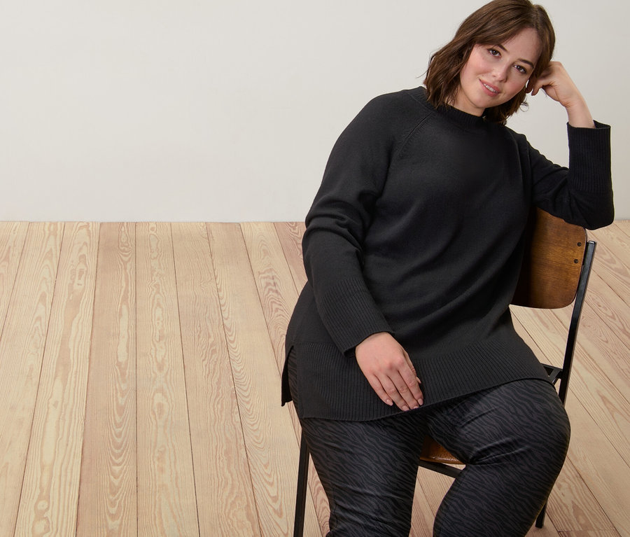 Une femme est assise sur une chaise et porte un long pull en maille noir et un pantalon imprimé en bengaline.