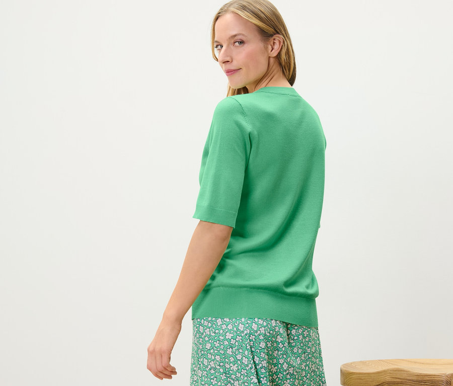 Une femme regarde par-dessus son épaule et porte un pull en maille fine vert et une jupe à fleurs.