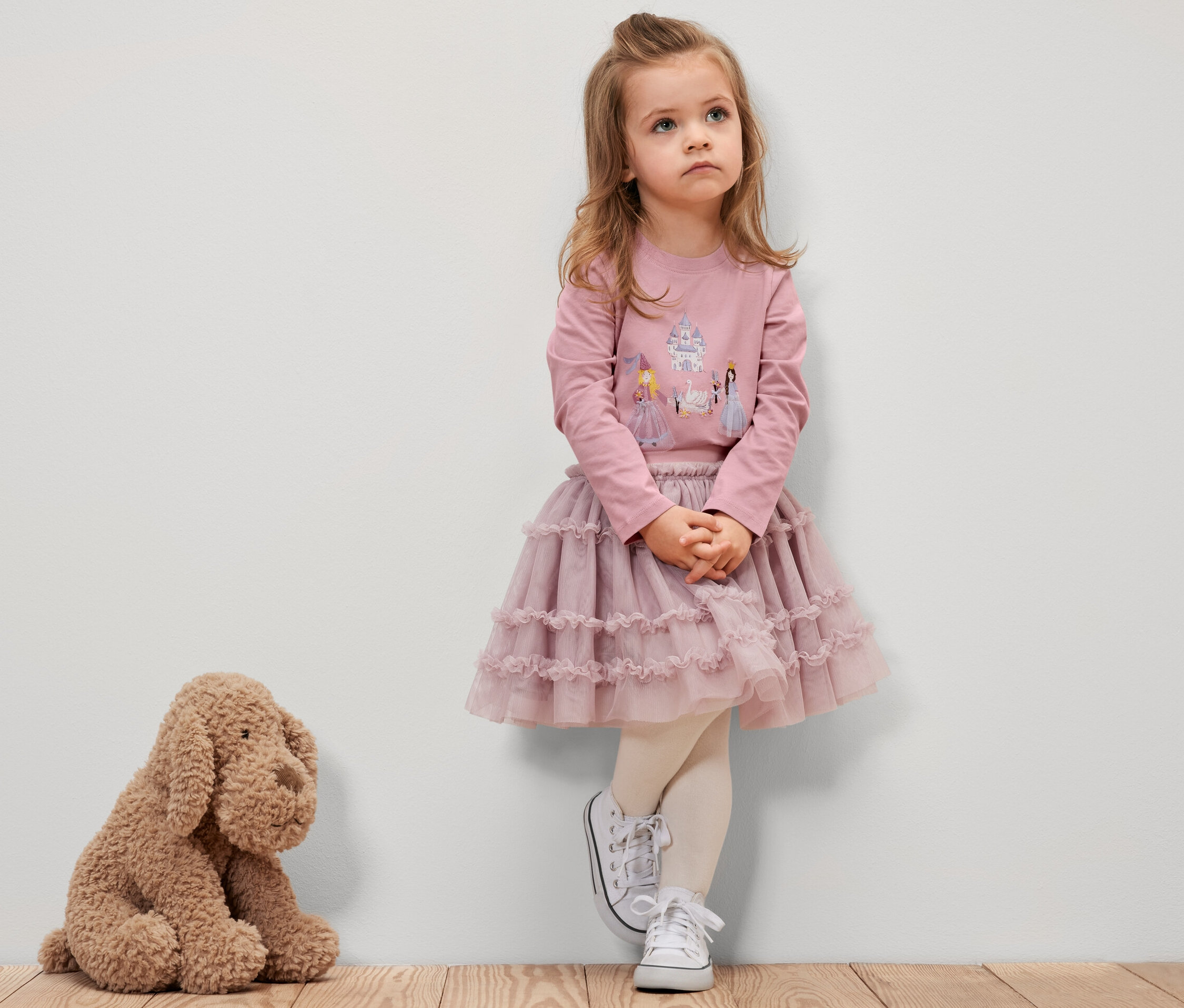 Une fillette vêtue d'un t-shirt rose à manches longues avec un motif de princesse, d'une jupe rose et de baskets blanches se tient contre un mur. Un chien en peluche est assis à côté d'elle.