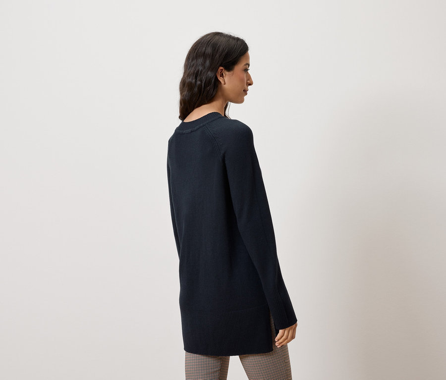 Une femme se tient de côté, portant un pull bleu foncé et un pantalon à carreaux.