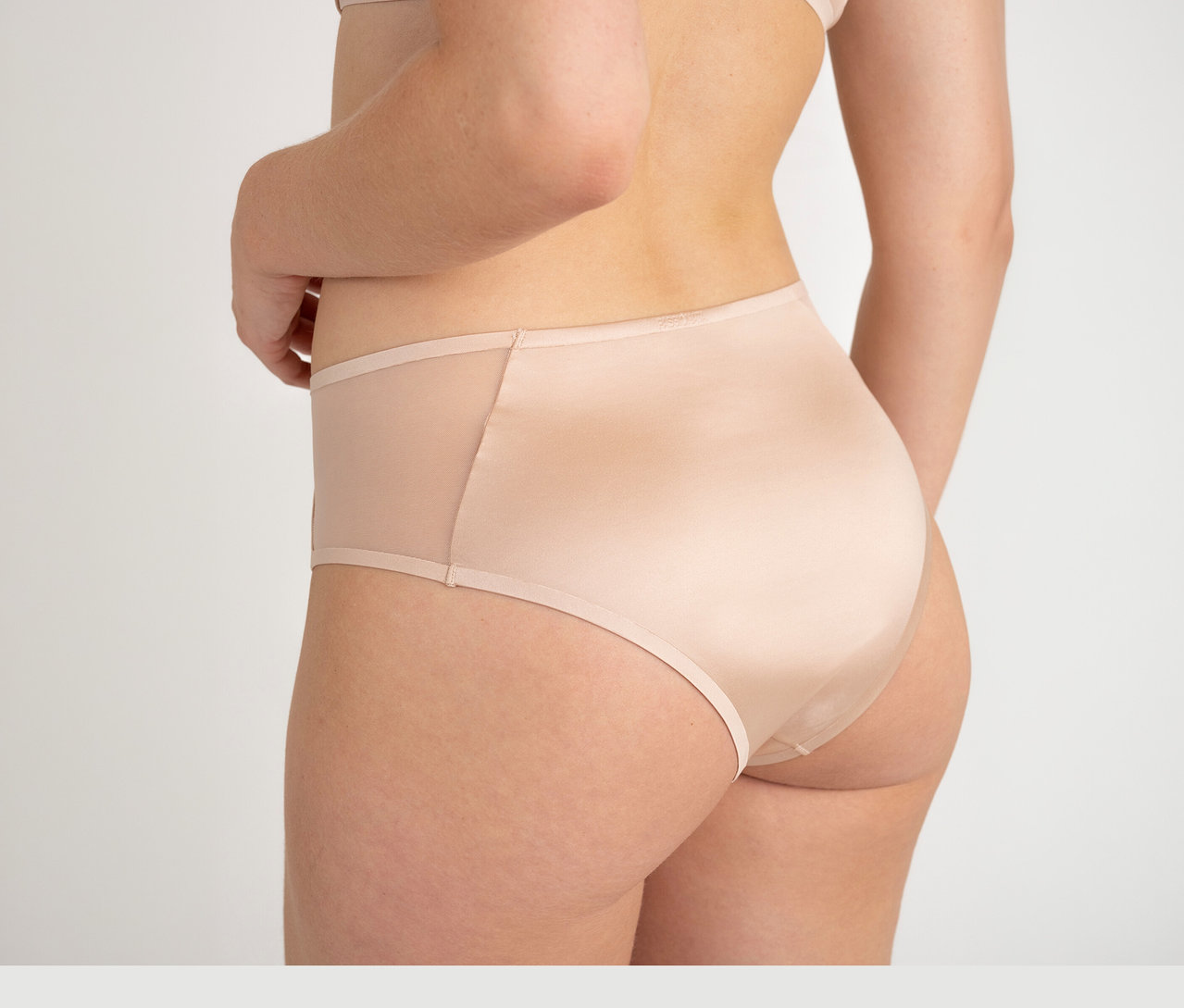 Une femme de dos porte un slip taille haute Passionata « Bianca » couleur dune avec des empiècements latéraux transparents.
