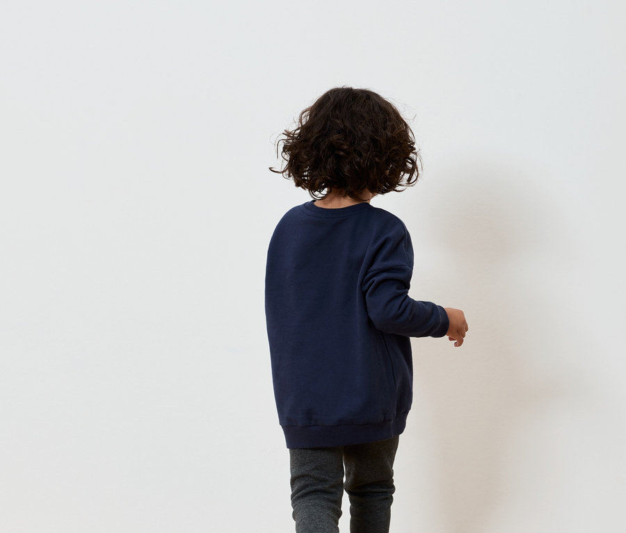 Un garçon aux cheveux bruns bouclés, portant un sweat-shirt bleu marine et un pantalon gris, se tient dos à un mur blanc.