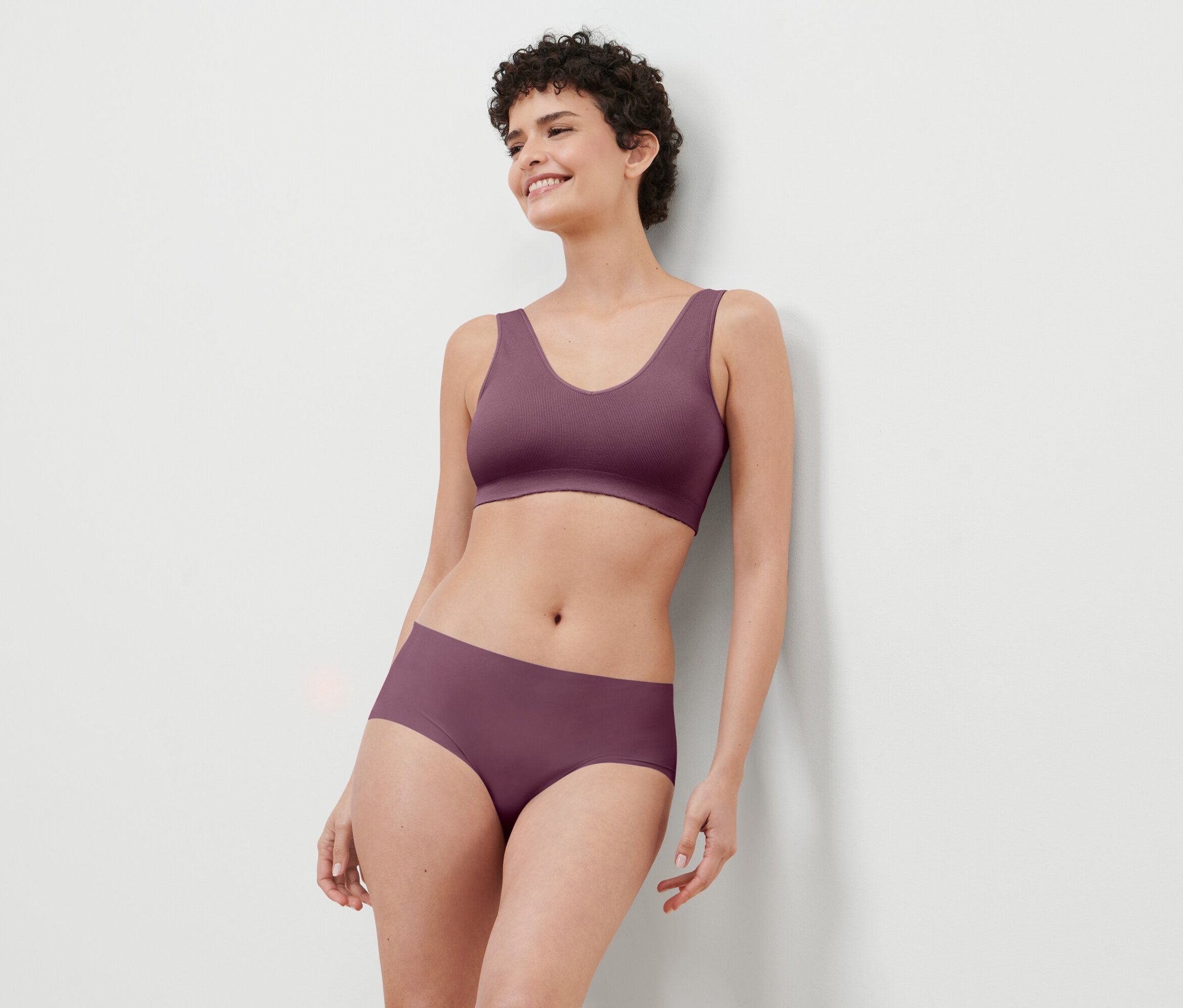 Une femme aux cheveux courts, portant un ensemble de lingerie bordeaux, est appuyée contre un mur blanc et sourit.