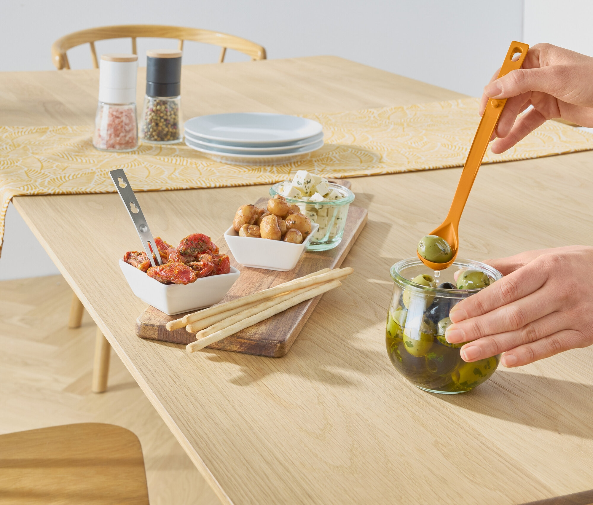 Une main prend des olives vertes dans un bocal avec une cuillère orange sur une table en bois. Sur la table, il y a des bols avec des tomates, des champignons, du fromage, des bâtonnets, des moulins à sel et à poivre.