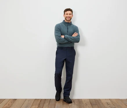 Un homme se tient debout, les bras croisés, portant un pull bleu-vert et un pantalon bleu foncé.