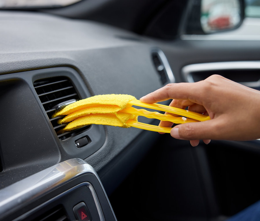 Une main nettoie une ventilation de voiture avec un plumeau jaune.