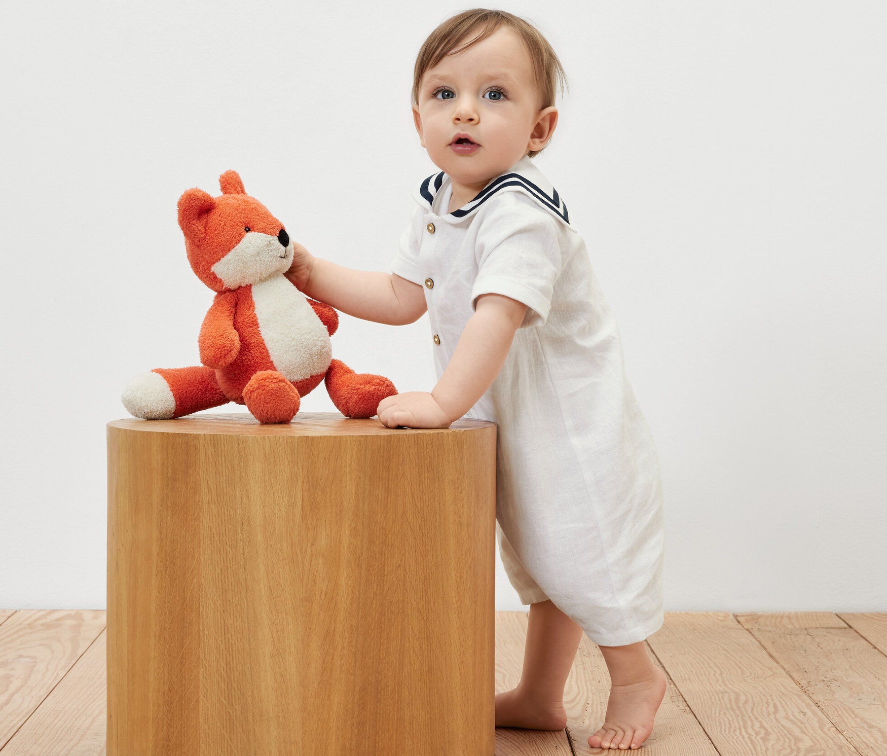 Un bébé en barboteuse de style marin s'appuie sur une table en bois et tient un renard en peluche.