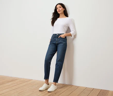 Une femme en chemisier blanc et jean bleu se tient contre un mur blanc.