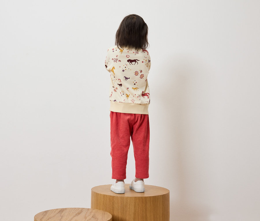 Un enfant, dos à la caméra, se tient sur un socle en bois, portant un sweat-shirt crème avec un imprimé de chevaux et un pantalon en velours côtelé rouge.