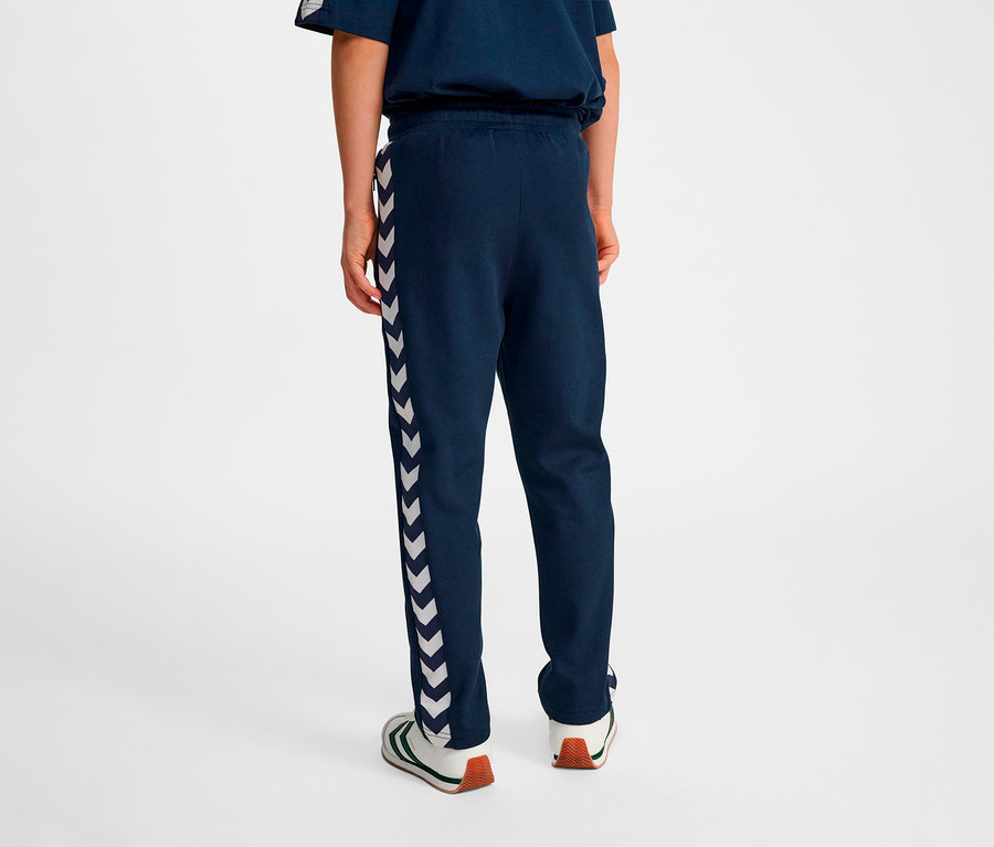 Personne vue de dos portant un pantalon bleu HUMMEL HMLJR Archive Regular Poly Pants avec des chevrons blancs sur le côté.
