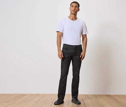 Un homme debout sur un plancher en bois, portant un jean noir et un t-shirt blanc.