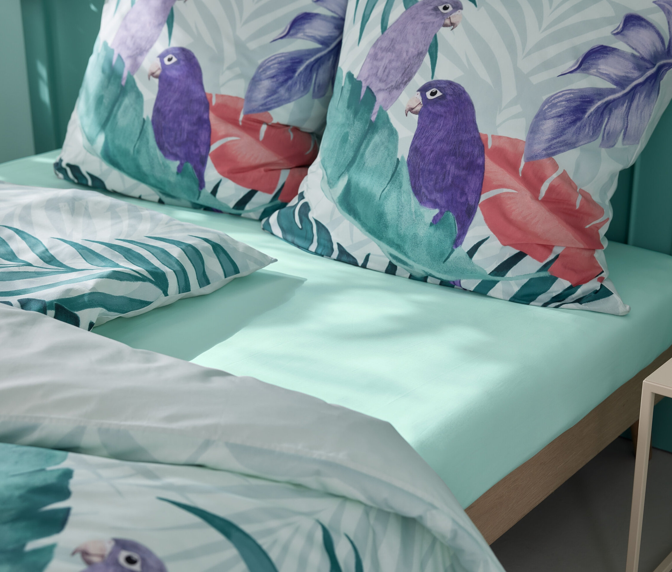 Drap-housse vert clair avec deux oreillers à motifs de perroquets et de feuilles tropicales.